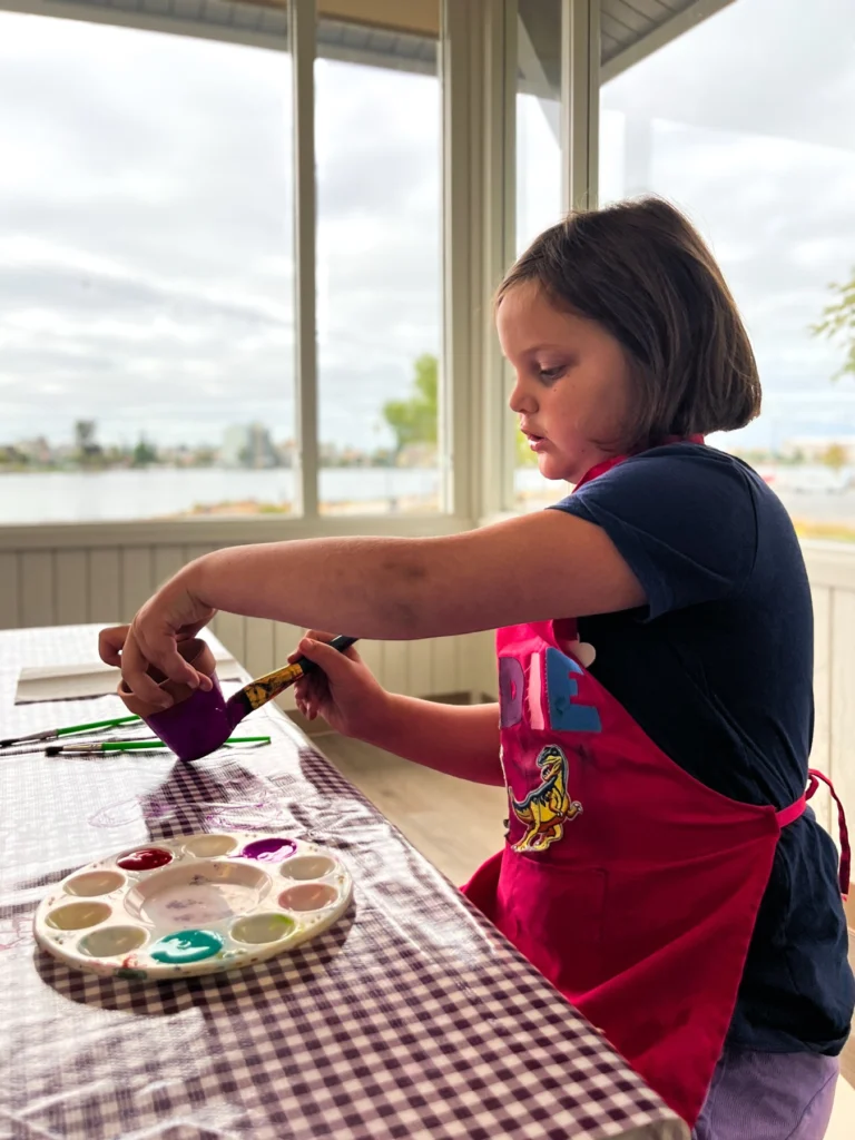 Camper in a red apron painting at a table by the window